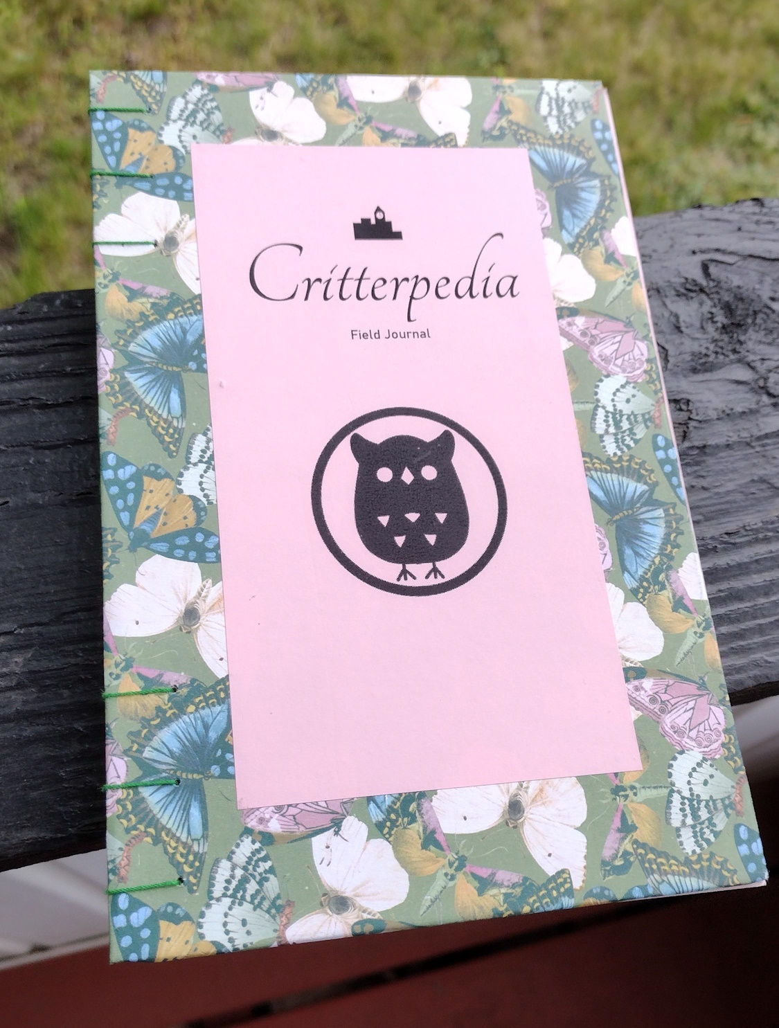 A hand bound journal. The covers are wrapped in green butterfly print paper. The top cover has a pink center with the words Critterpedia Field Journal as the title. Above this title is a small icon of a city skyline. Below the title is a large owl icon, based on the museum icon