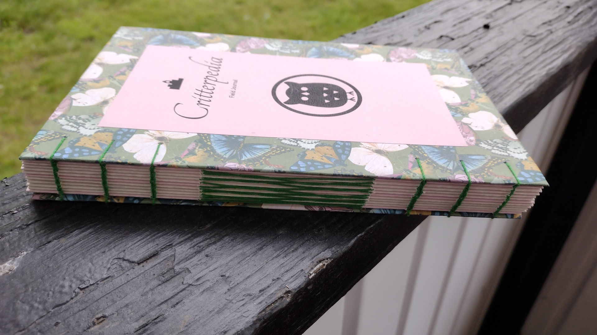 The spine of the book. The folded edges of the pages are still visible between the covers. It's held together with green sewing. On the two ends are three lines each of coptic style chain stitching. In the center is a section of french link, forming an intertwined ladder.